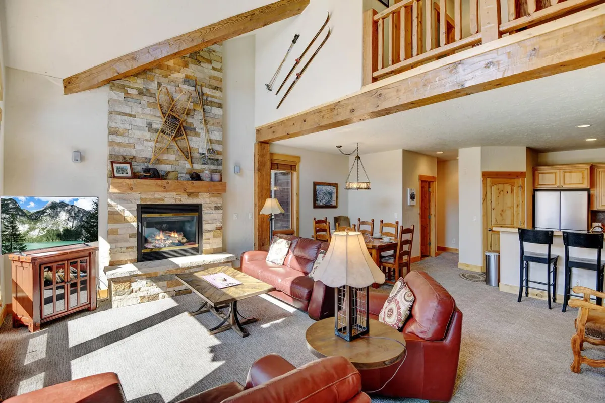This image features a spacious great room with a stone fireplace, plush leather couches, a large dining area, and expansive windows that showcase beautiful mountain views. The wall-mounted TV is visible, anchored by the warmth of the fire, while the open-plan layout lets natural light flood every corner.