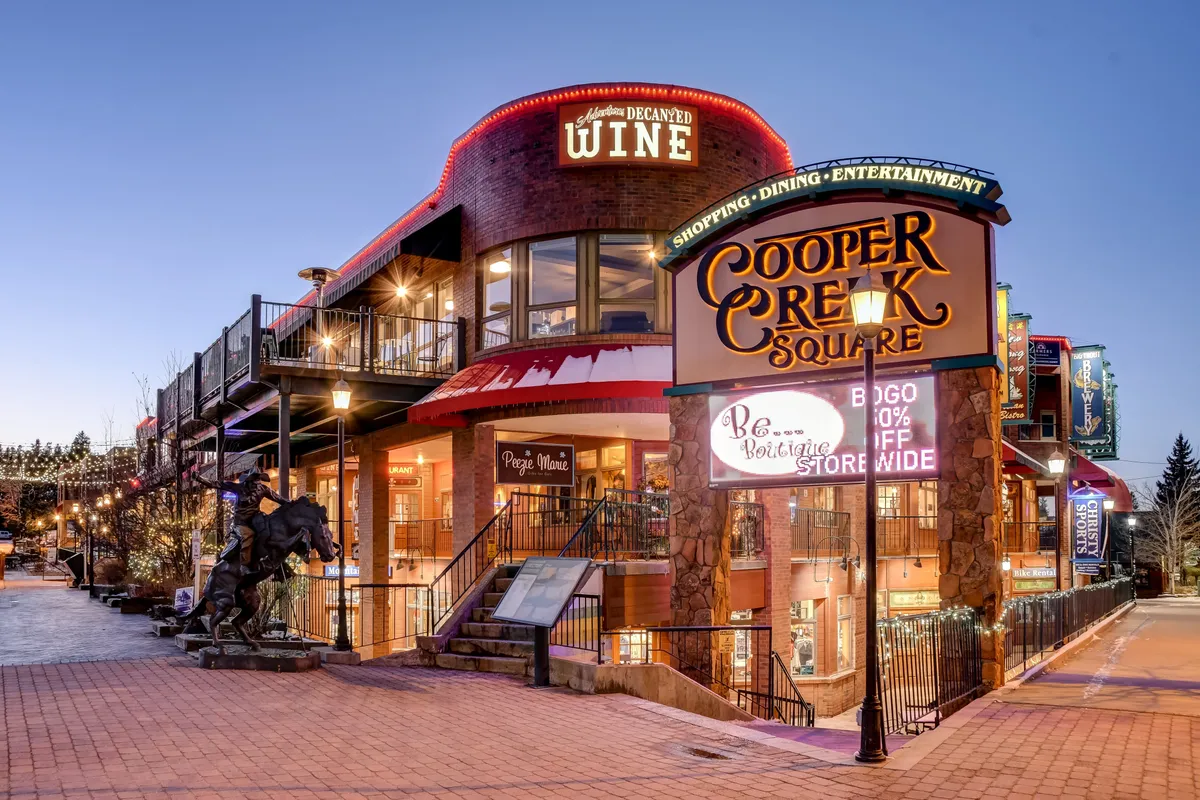 A festive exterior view of Cooper Creek Square, featuring signage for shopping and dining, a staircase to an upper level, and a public sculpture. The setting is lively, with hints of social gatherings, wine tastings, and holiday celebrations.
