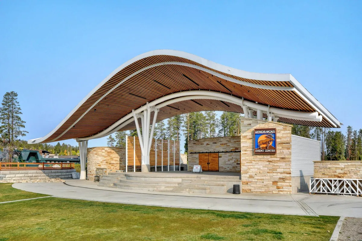 An exterior shot of a modern curved-roof structure with a stone wall and steps leading up to a stage, set against an inviting backdrop. The venue suggests a lively gathering place for community events, performances, and celebrations, with an open-air, energetic atmosphere.