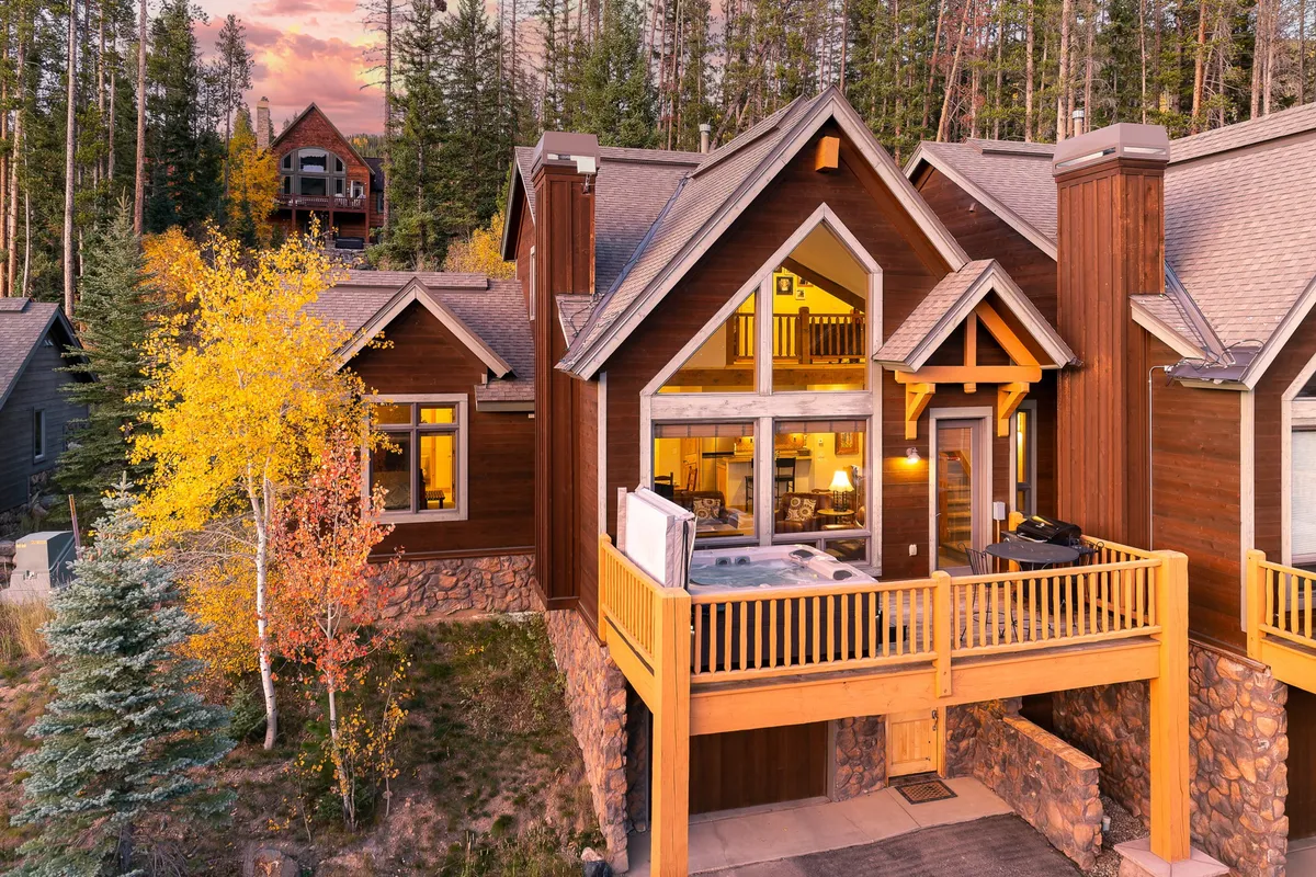 The image showcases the exterior of a luxurious mountain villa, featuring a spacious wooden balcony with ample space for relaxation or gatherings. Large windows frame the home, offering glimpses of natural light and inviting the scenic forested surroundings indoors. The sturdy stone foundation and the backdrop of towering pines create a cozy, inviting atmosphere, perfect for both summer barbecues and winter retreats.