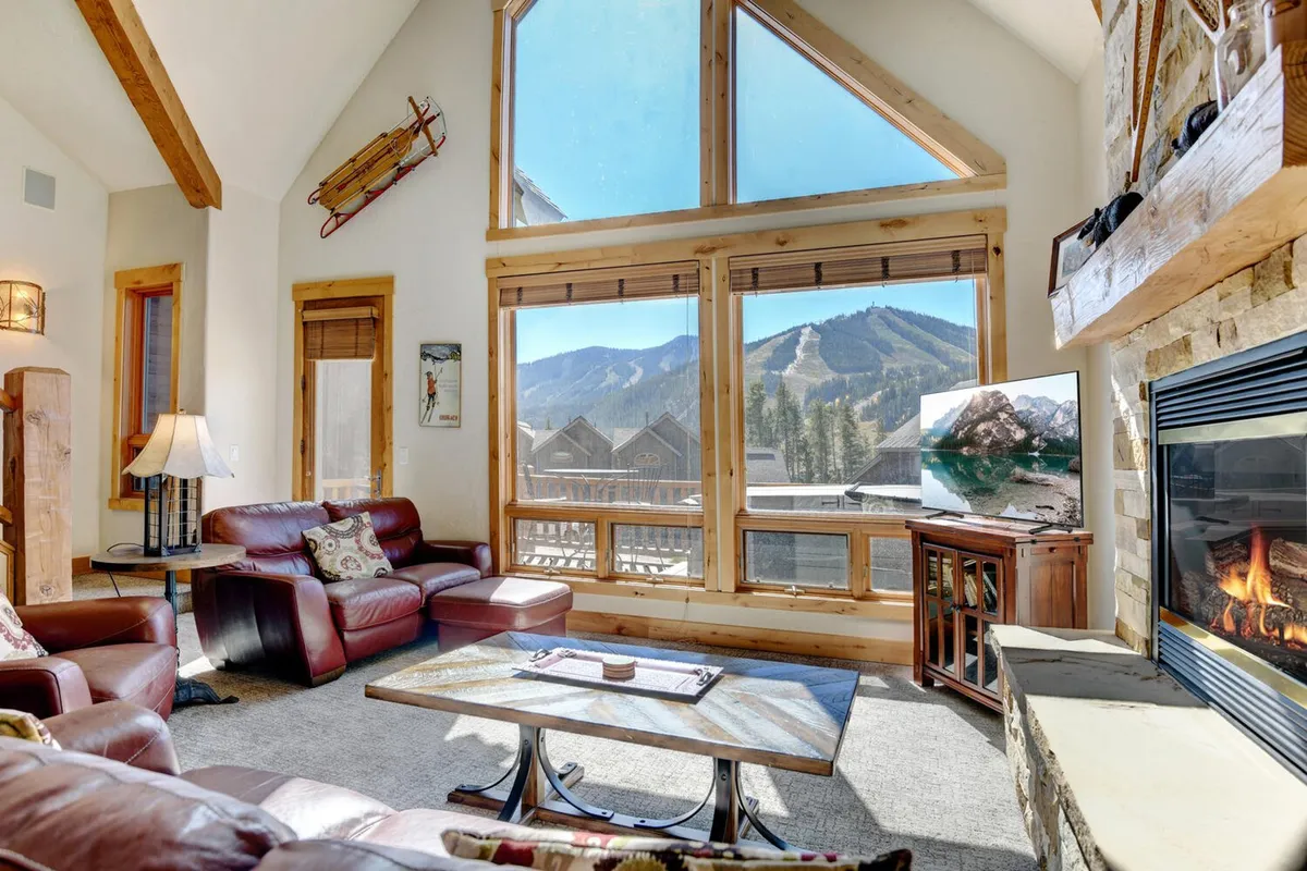 An inviting living room with a cozy fireplace, leather seating, a coffee table, and expansive windows overlooking the mountains. The space feels equally suited for shared stories by the fire after a day outdoors or enjoying morning coffee while watching the sunrise over the peaks.