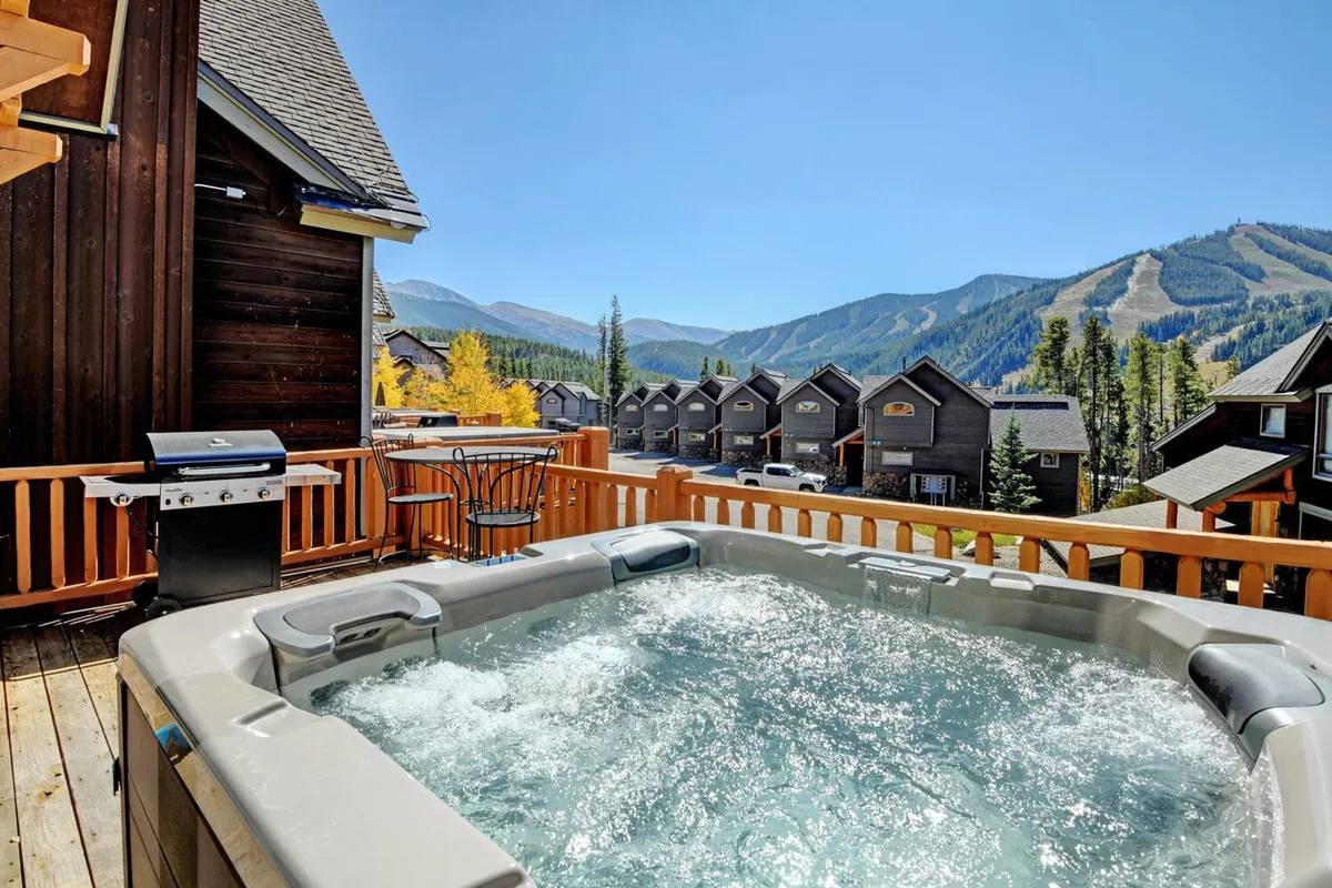 This image showcases the hot tub in the foreground, the deck area with a BBQ grill, and abundant mountain scenery. The soft lighting suggests evening relaxation, while the setup invites guests to imagine socializing or savoring an alfresco meal outdoors.