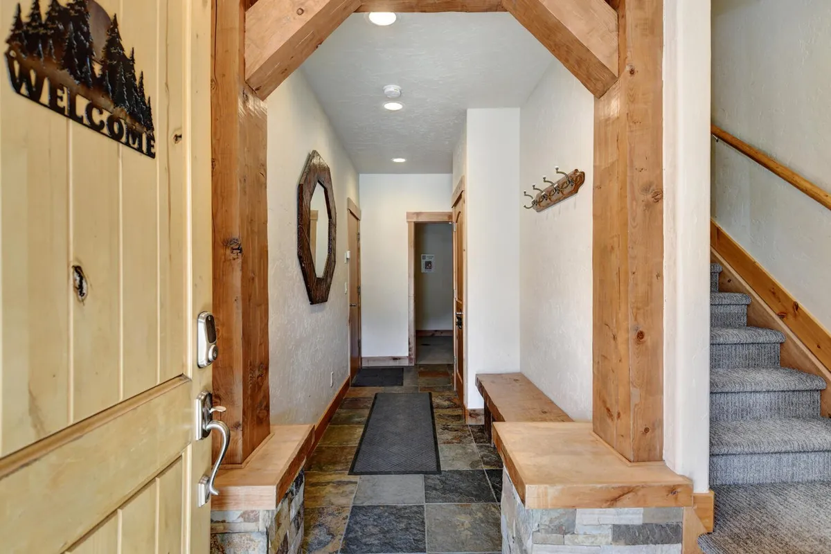 This image showcases the home's inviting entryway, featuring a wooden front door with a 'WELCOME' sign, an arched wooden entryway, a large mirror, a stone bench perfect for taking off boots or setting down bags, and a staircase leading upstairs. The scene is warm, organized, and perfectly suited for a mountain home.