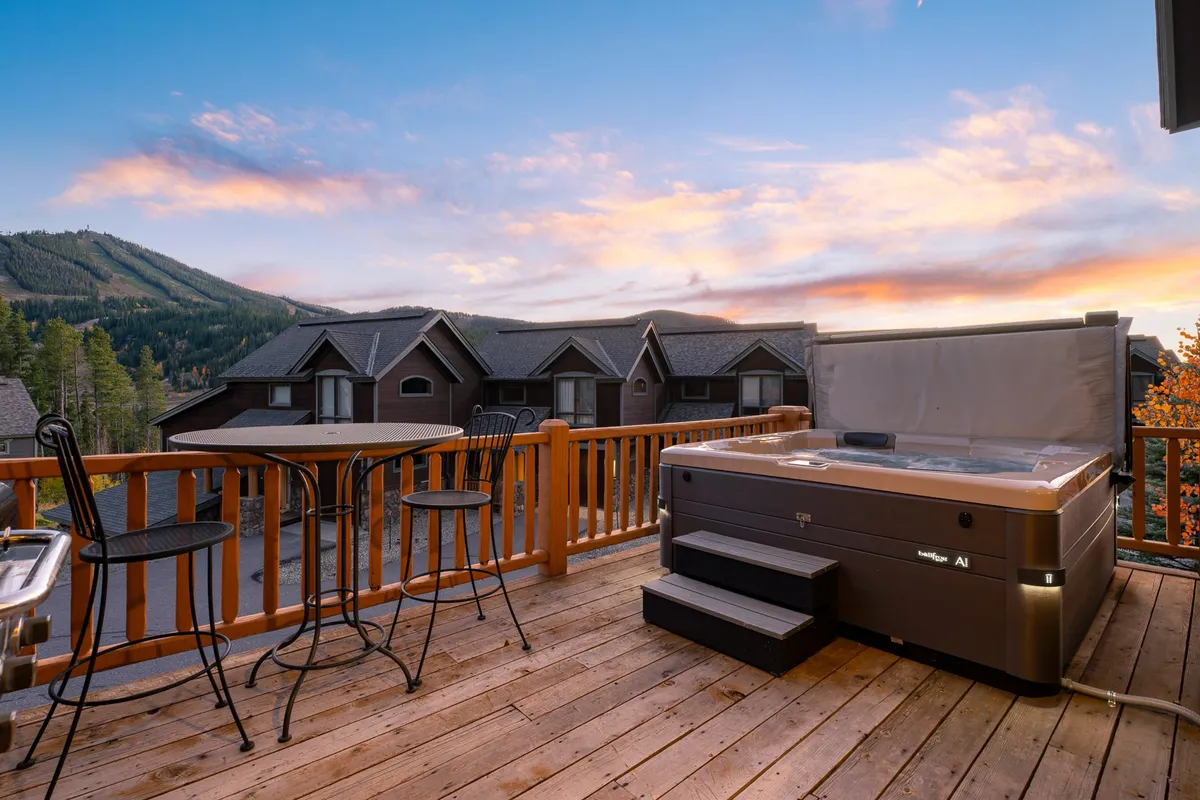 This exterior shot presents the hot tub and outdoor seating area from a slightly different angle, emphasizing the spaciousness of the deck and the unobstructed panoramic views of the mountains during a sunset sky. Friends are gathered, suggesting togetherness and shared enjoyment.