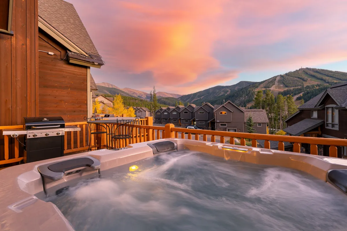 This exterior image captures a private deck featuring a brand new hot tub with bubbling water, a BBQ grill to the side, comfortable outdoor seating, and a stunning view of the snow-capped mountains at sunset. The inviting scene suggests relaxation and wellness, pairing both the luxury of the hot tub and the allure of mountain vistas.