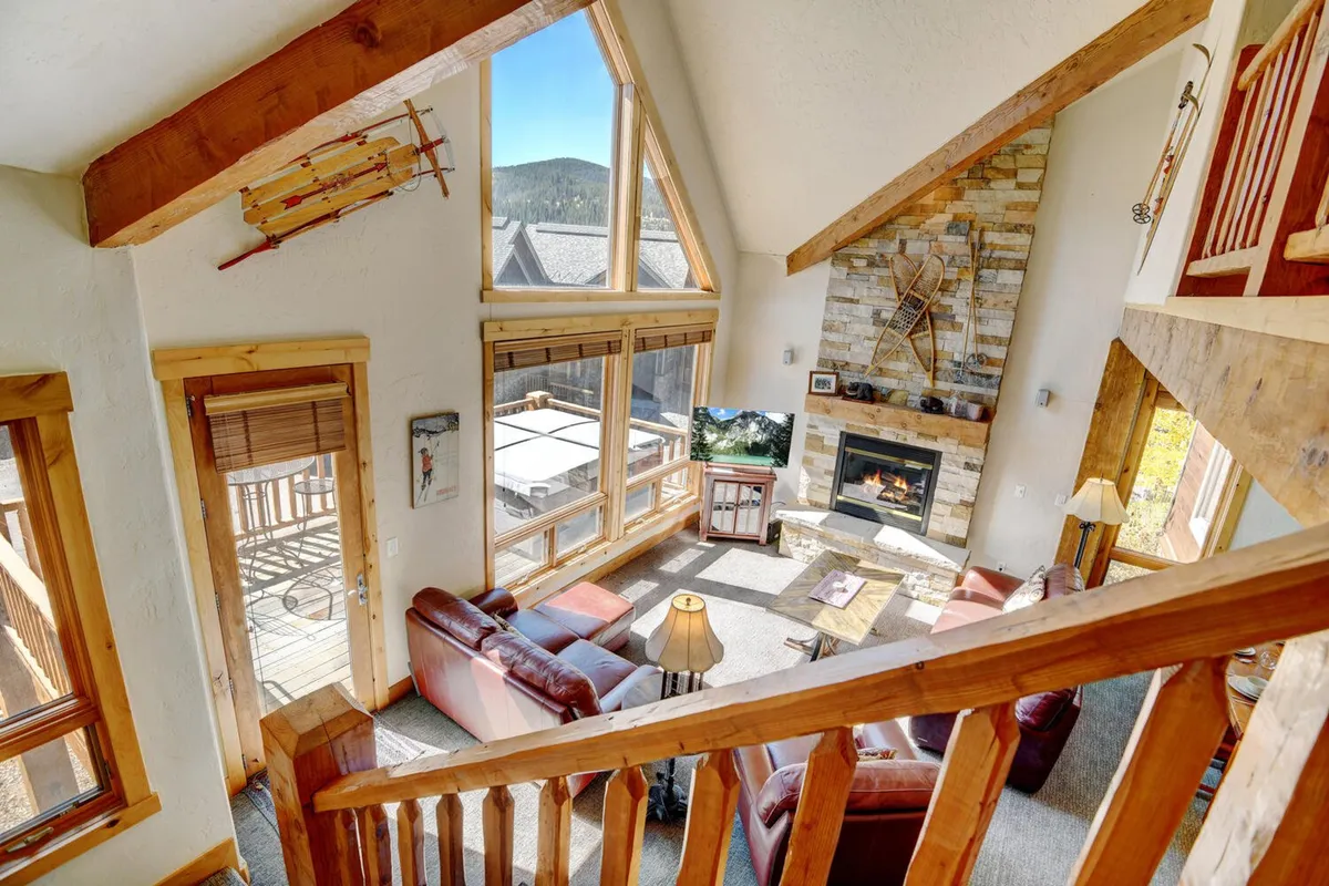 A spacious living area with a stone fireplace, exposed wooden beams, and large windows framing mountain views. Plush seating invites relaxation, whether gathering by the fire in winter or soaking up sunlight and scenery during summer. The scene embodies comfort paired with a strong connection to the outdoors.