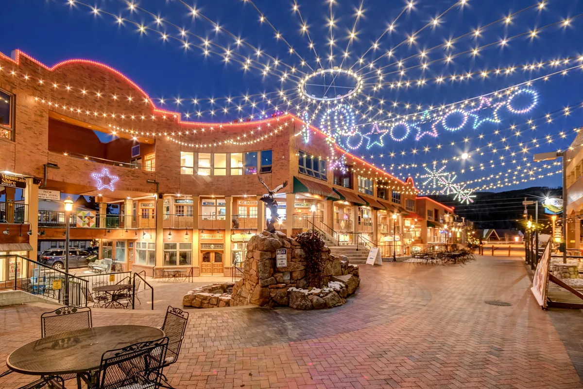 Twinkling lights hang overhead, illuminating the outdoor stone fountain and a cozy seating area, suggesting evening relaxation or social gatherings in a magical mountain setting.